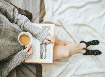 woman holding a cup of coffee at right hand and reading book on her lap while holding it open with her left hand in a well-lit room